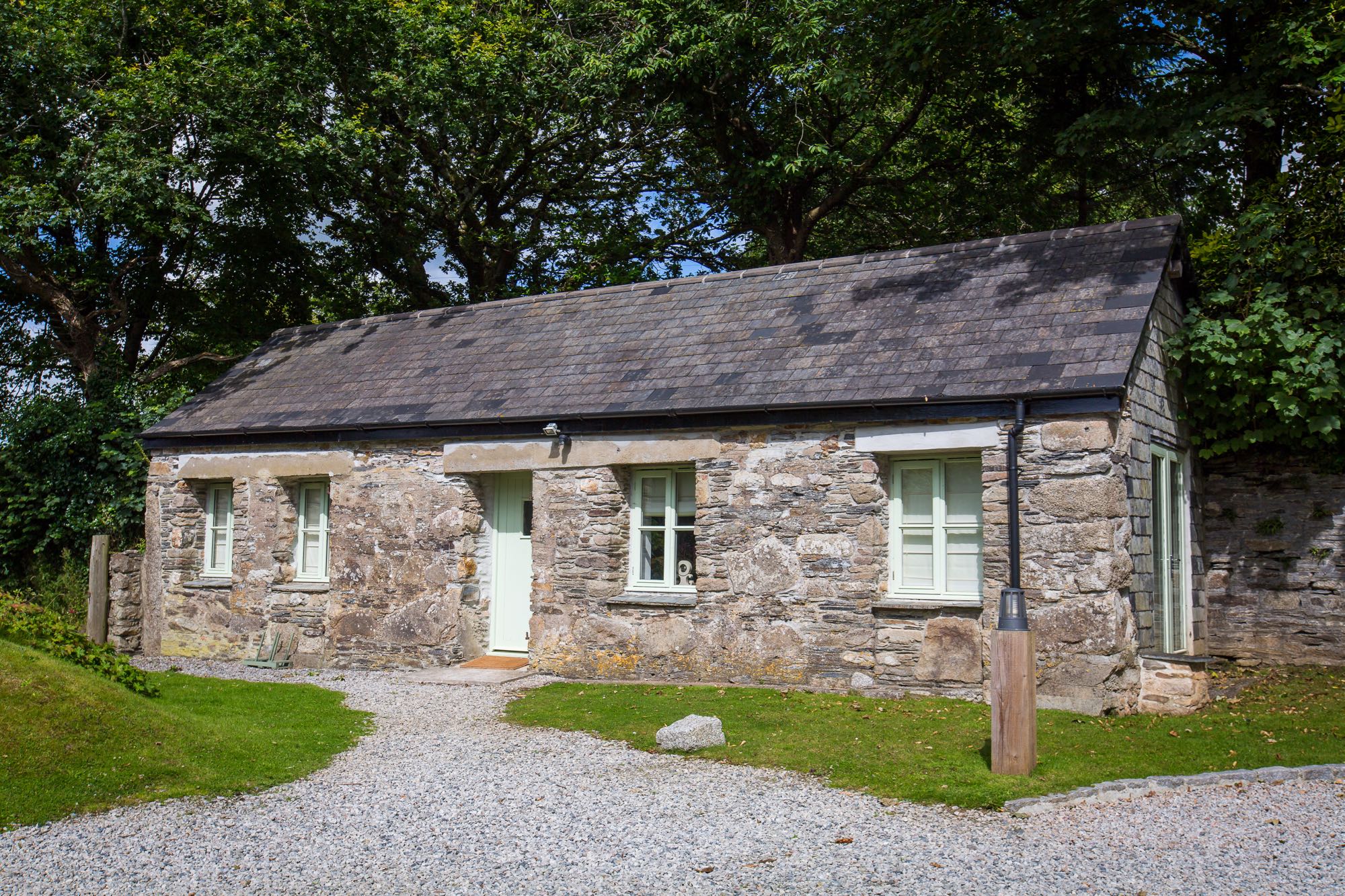 This is a photo of cottage number one. Cottage number one sleeps two people. The proerty is single storey. The photo shows the full width of the cottage. In the background you can see trees which are in leaf. In the foreground you can see two patches of grass one on the left and one on the right and in the front there is gravel. The cottage is made of granite stone and is brown and grey in colour. The roof is constructed of grey slate and is pitched with the roof line running from left to right of the photograph along the length of the cottage. In the middle of the front of the cottage there is a wooden door that is painted pale green on either side of the story there are two windows on each side of this door. They are georgian style sash windows and also painted green. On the right hand side of the cottage the gable end has hanging slate on it. There is a French window in this end of the cottage and it is also painted green. In the foreground of the picture there is a light bollard for outside lighting.