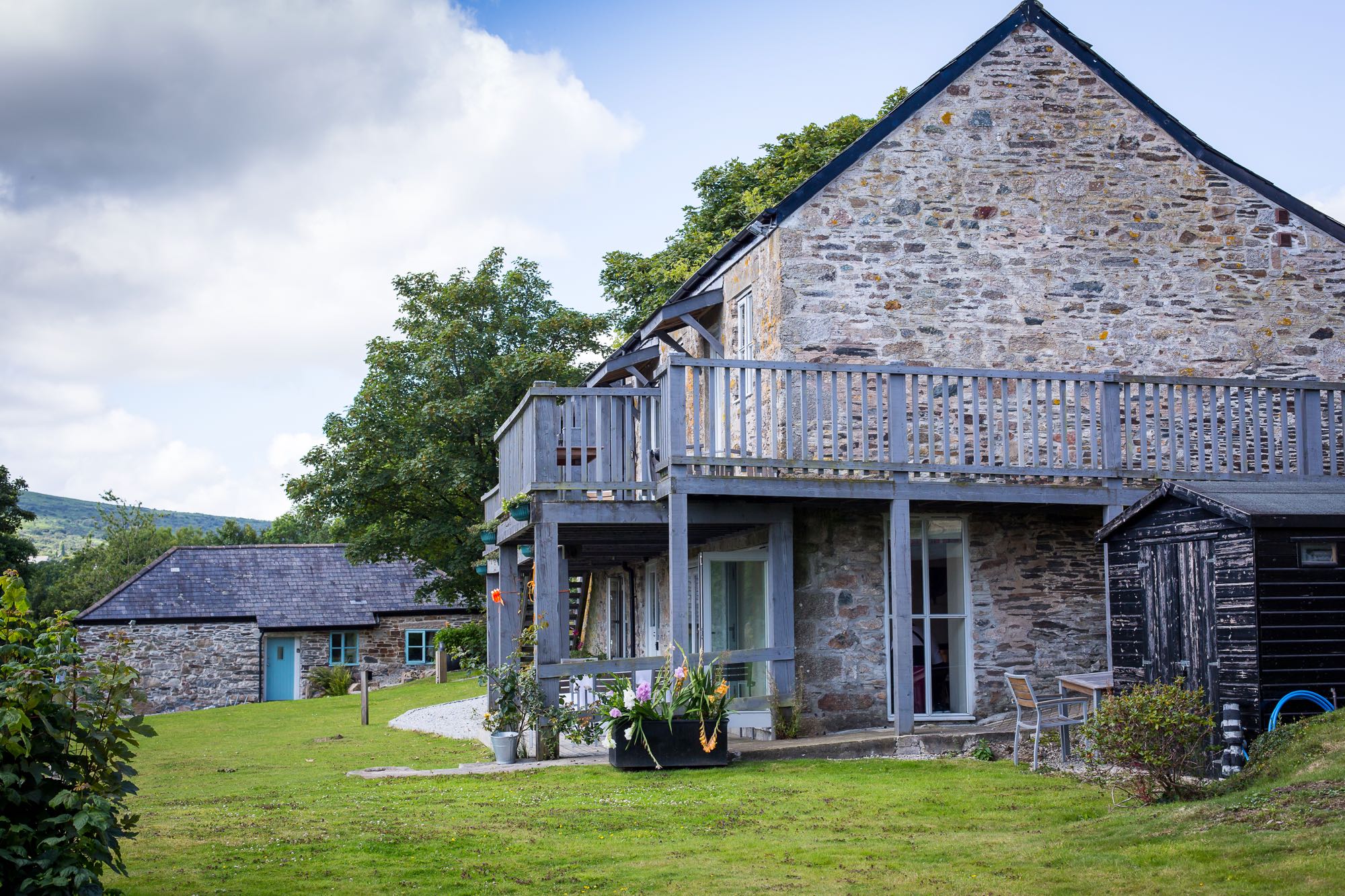 This is an exterior shot of a large barn that is split into three cottages namely cottage three, cottage four and cottage five. The barn has two storeys. Cottage three and cottage four are upstairs. Cottage five is on the ground floor. In the image the large barn is seen from the side. The side of the barn is made of local granite cob in gray and brown. It has a pitched roof with a gable end. There is a window on the bottom left hand corner. This is a window of cottage 5. The window is a four paned casement style window and is painted pale gray. To the front of the building (which is on the left side of the photograph) there is a set of grey painted french windows (these lead into cottage 5). There is a balcony along the full length of the second floor. The balcony is painted grey and is made of wood. The balcony is supported on wooden posts from the ground. The area immediately around the cottage is mostly lawn with a gravel path in the perimeter immediately around the cottage. In the left hand corner of the picture there is a hill sloping gently off to the left. Cottage number 2 can be sen in the backgound. This is a detached cob cottgae with gray slate roof and a blue painted door and windows. In the backgound there are some trees leaf. The sky is overcast.